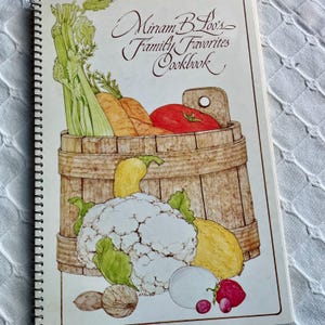 May include: A spiral-bound cookbook titled Miriam B. Loo's Family Favorites Cookbook. The cover features a watercolor illustration of a wooden basket filled with celery, carrots, and a tomato, with cauliflower, squash, and berries below.