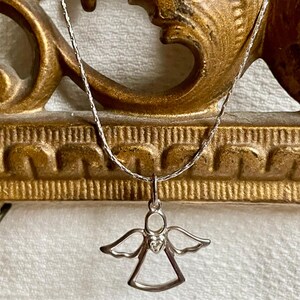 K&ouml;nnte beinhalten: Eine silberfarbene Halskette mit einem Anh&auml;nger in Form eines Engels. Der Engel-Anh&auml;nger hat ausgebreitete Fl&uuml;gel und ein herzf&ouml;rmiges Detail. Die Halskette wird vor einem goldfarbenen, verzierten Hintergrund pr&auml;sentiert.