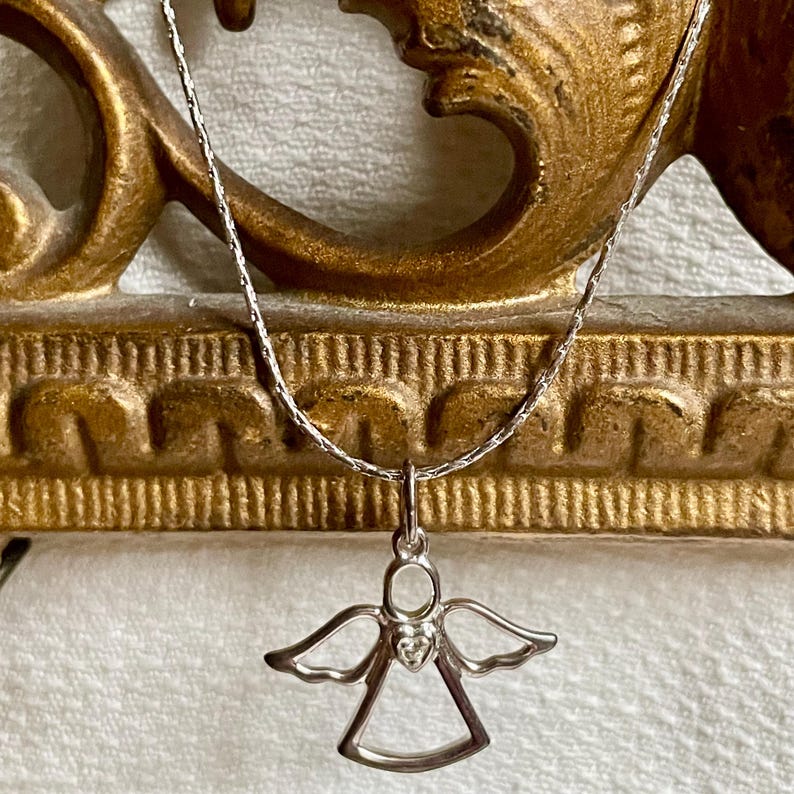 K&ouml;nnte beinhalten: Eine silberfarbene Halskette mit einem Anh&auml;nger in Form eines Engels. Der Engel-Anh&auml;nger hat ausgebreitete Fl&uuml;gel und ein herzf&ouml;rmiges Detail. Die Halskette wird vor einem goldfarbenen, verzierten Hintergrund pr&auml;sentiert.