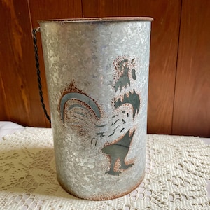 May include: A cylindrical galvanized metal container featuring a cutout silhouette of a rooster. The surface has a mottled gray finish with rust-colored accents along the edges and the rooster cutout. A twisted metal handle is attached to the side.