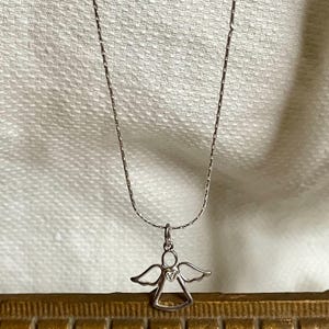 K&ouml;nnte beinhalten: Eine silberfarbene Halskette mit einem kleinen Anh&auml;nger in Form eines Engels. Der Anh&auml;nger hat ausgebreitete Fl&uuml;gel und ein herzf&ouml;rmiges Detail. Die Halskette h&auml;ngt vor einem weichen, cremefarbenen Hintergrund.