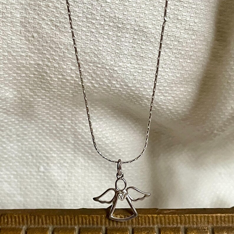 K&ouml;nnte beinhalten: Eine silberfarbene Halskette mit einem kleinen Anh&auml;nger in Form eines Engels. Der Anh&auml;nger hat ausgebreitete Fl&uuml;gel und ein herzf&ouml;rmiges Detail. Die Halskette h&auml;ngt vor einem weichen, cremefarbenen Hintergrund.