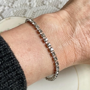May include: A silver-toned bracelet with a line of clear, square-cut rhinestones. The bracelet is worn on a wrist, and the clasp is visible. The bracelet is a delicate piece of jewelry.