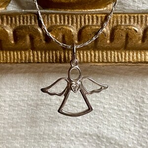 K&ouml;nnte beinhalten: Ein silberner Anh&auml;nger in Form eines Engels mit einem kleinen herzf&ouml;rmigen Edelstein in der Mitte. Der Anh&auml;nger h&auml;ngt an einer filigranen Silberkette. Der Engel hat ausgebreitete Fl&uuml;gel und einen Heiligenschein. Der Hintergrund ist eine strukturierte, cremefarbene Oberfl&auml;che.