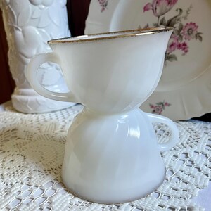 May include: Two stacked white milk glass teacups featuring a swirling ribbed pattern and a thin gold rim on the top cup. Each cup has a curved handle extending from the side. The cups are made of translucent white glass with a glossy finish.