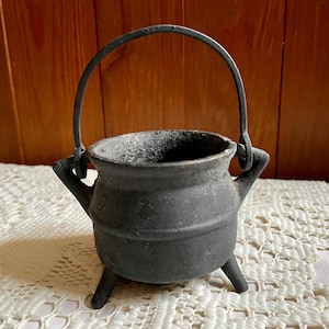 May include: A small, matte black cast iron cauldron featuring a rounded body, three short legs, and a curved metal handle attached to two side lugs. The surface has a textured, weathered appearance with a wide, open top rim.