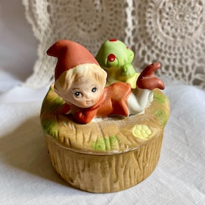 May include: A ceramic trinket box featuring a whimsical design. The box is shaped like a mushroom and has a detailed, textured exterior. A small figurine of a gnome wearing a red hat and outfit is on top, along with a green mushroom.