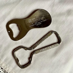 May include: Two vintage metal bottle openers. One is shaped like a teardrop with "The Raphad" and city names. The other is a loop with "Schmidt's of Philadelphia" engraved on it. Both are silver-toned.