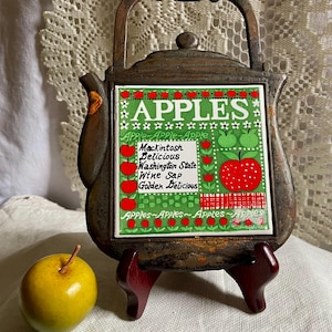 May include: A cast iron teapot-shaped trivet with a central ceramic tile featuring the text "APPLES" and a list of apple varieties including Mackintosh, Delicious, Washington State, Wine Sap, and Golden Delicious, accompanied by a yellow apple.
