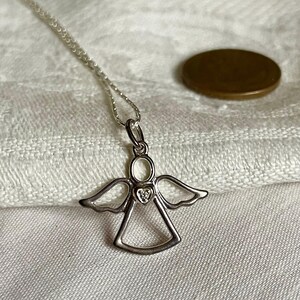 K&ouml;nnte beinhalten: Eine silberne Engelsanh&auml;nger-Halskette mit einem kleinen herzf&ouml;rmigen Edelstein in der Mitte. Der Engel hat ausgebreitete Fl&uuml;gel und einen Heiligenschein. Die Halskette liegt auf einer wei&szlig;en Stoffoberfl&auml;che, im Hintergrund eine M&uuml;nze.