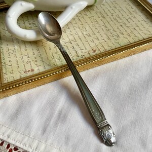 May include: A silver spoon with an ornate handle featuring a floral design at the base. The spoon is resting on a white surface, with a framed document in the background. The spoon's handle has a decorative pattern.
