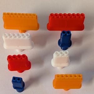May include: An assortment of colorful, plastic toy engine components in orange, red, white, and blue. The components include engine blocks and other parts, likely for a model car or construction set. The pieces are arranged on a white surface.