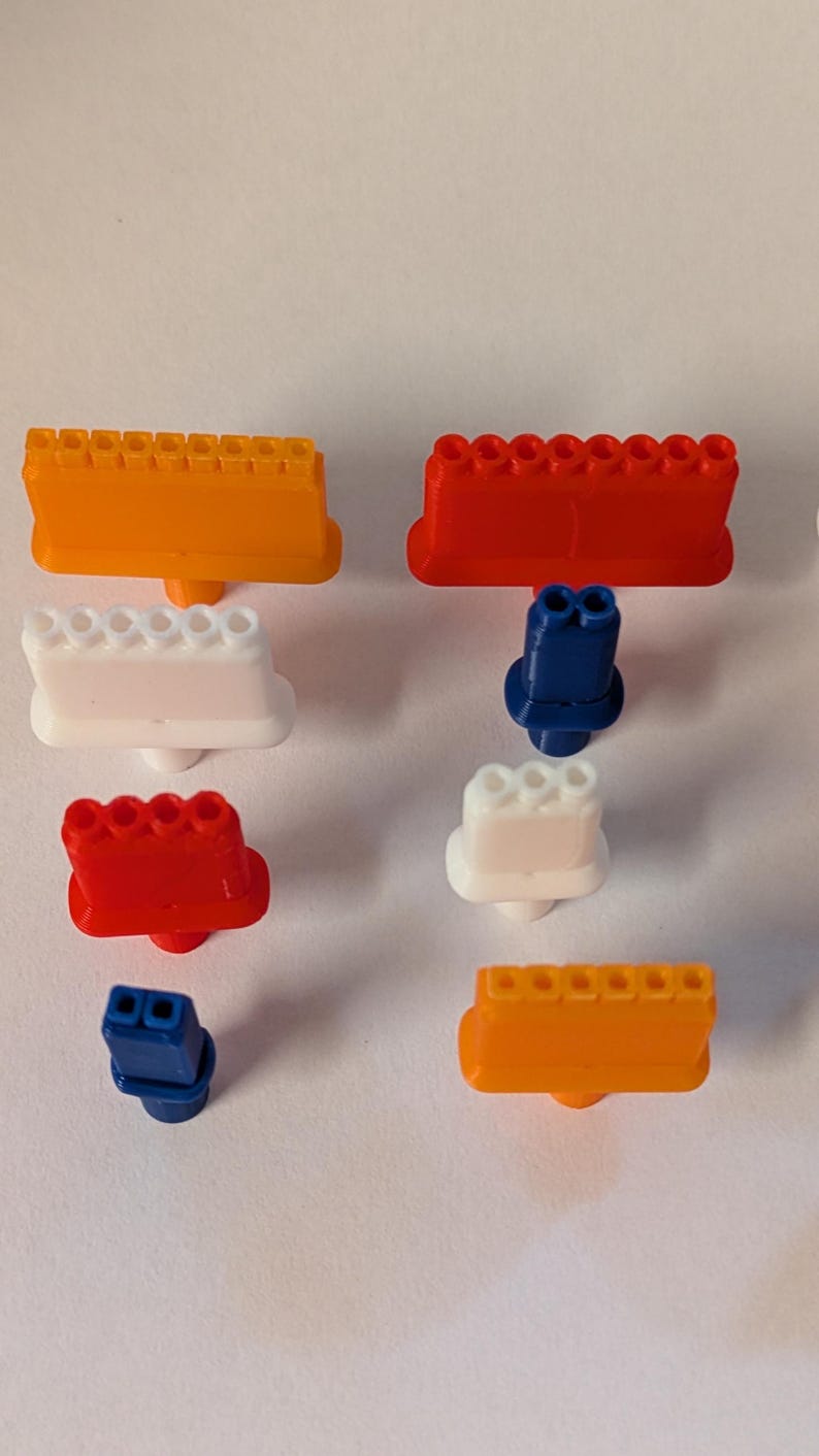 May include: An assortment of colorful, plastic toy engine components in orange, red, white, and blue. The components include engine blocks and other parts, likely for a model car or construction set. The pieces are arranged on a white surface.