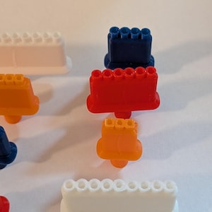 May include: An assortment of colorful, plastic toy building blocks in various shapes and sizes. The blocks are in shades of red, orange, blue, and white. The image is taken from a high angle, with the blocks scattered on a white surface.