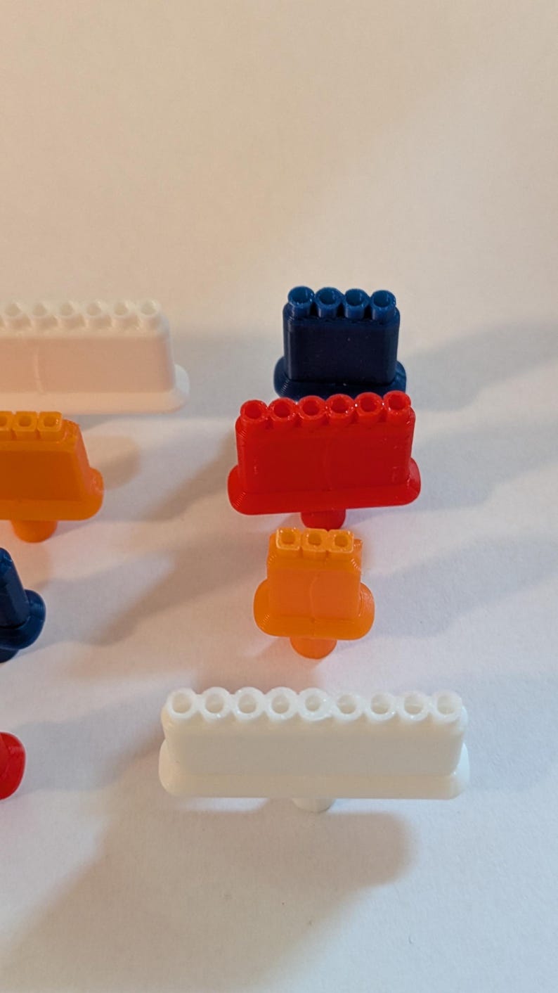 May include: An assortment of colorful, plastic toy building blocks in various shapes and sizes. The blocks are in shades of red, orange, blue, and white. The image is taken from a high angle, with the blocks scattered on a white surface.