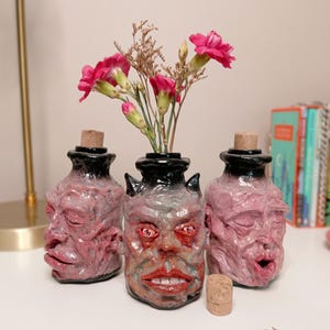May include: Three ceramic bottles shaped like human heads with pinkish-red skin tones and black glazed tops. The central bottle features a wide-eyed expression with visible teeth and holds a small bouquet of pink carnations and dried stems.