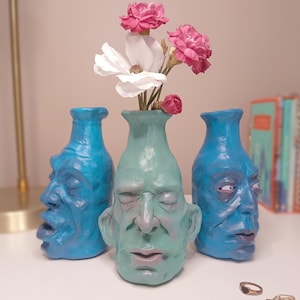May include: Three ceramic vases shaped like human heads with facial features. The two outer vases are bright blue, while the centre vase is a pale mint green. The centre vase holds a small bouquet of white and pink flowers, with visible petals.