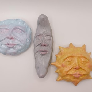 May include: Three wall-mounted sculptures featuring celestial faces with closed eyes. A round light blue full moon, a tall grey crescent moon, and a yellow sun with pointed rays. Each piece has a textured surface and subtle pink accents.