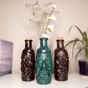 May include: Three glass bottles with sculpted human face designs on the exterior. Two bottles are dark brown and one central bottle is teal green. Each bottle is topped with a light brown cork stopper. The centre bottle holds a sprig of dried flowers.