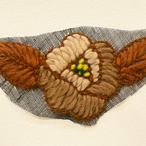May include: A handmade textile flower with brown and tan yarn petals and leaves, and a center with yellow and green accents. The flower is stitched onto a piece of black and white fabric, set against a white background.