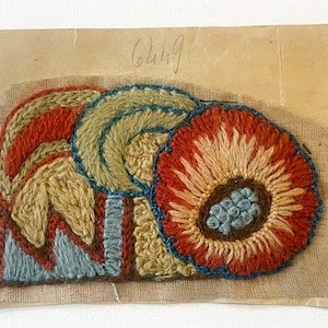 May include: A colorful, hand-stitched textile art piece featuring floral and geometric designs. The piece showcases a large red and orange flower with a blue center, alongside other abstract shapes in shades of yellow, blue, and red. The number "6449" is handwritten at the top.