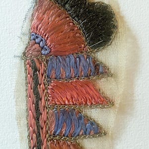 May include: Close-up of an embroidered textile with a layered design. The piece features shades of red, blue, and black thread, outlined with metallic gold thread. The textile is mounted on a cream-colored fabric.