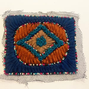 May include: A rectangular textile art piece with a central oval design in shades of brown and teal, surrounded by a blue border. The design is embellished with beads and a black mesh border.