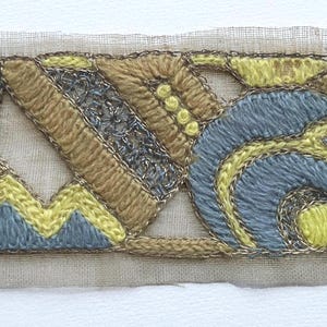 May include: Embroidered fabric patch featuring a geometric design in shades of blue, yellow, and tan. The embroidery includes a metallic silver thread outline, creating a decorative border. The patch is mounted on a sheer, off-white fabric.