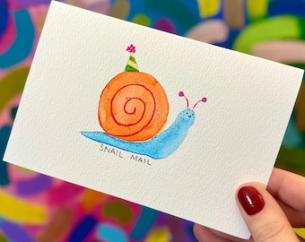 May include: A white card with a blue snail wearing a party hat and carrying a spiral orange shell. The text "Snail Mail" is written below the snail.