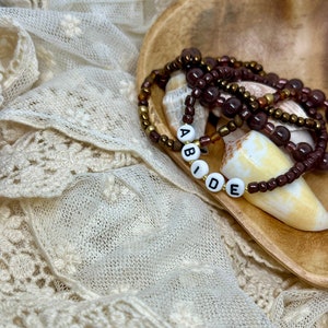 May include: A close-up of a beaded bracelet with the word "ABIDE" spelled out in white letters. The bracelet is made of brown and gold beads and is resting on a wooden surface with a white lace fabric background.