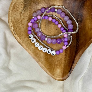 May include: A set of four purple and silver beaded bracelets with the word "BESTILL" spelled out in white letters.