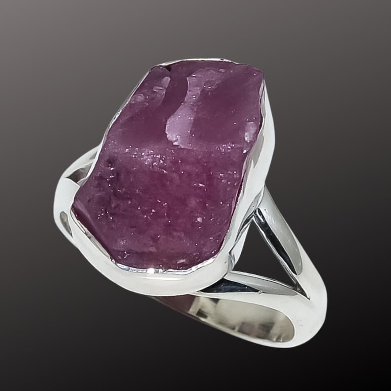 K&ouml;nnte beinhalten: Ein Silberring mit einem gro&szlig;en, roh geschnittenen, purpurrosa Edelstein. Der Stein ist in einer L&uuml;nettenfassung gefasst, und der Ring hat ein geteiltes Schaftdesign. Der Edelstein hat ein nat&uuml;rliches, strukturiertes Aussehen.