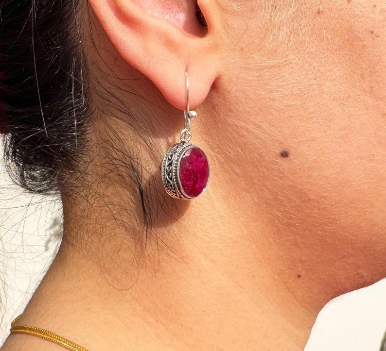 Op de afbeelding: Zilveren oorhangers met een ronde, rode edelsteen in een zilveren zetting. De oorbellen hebben een delicaat, versierd ontwerp.