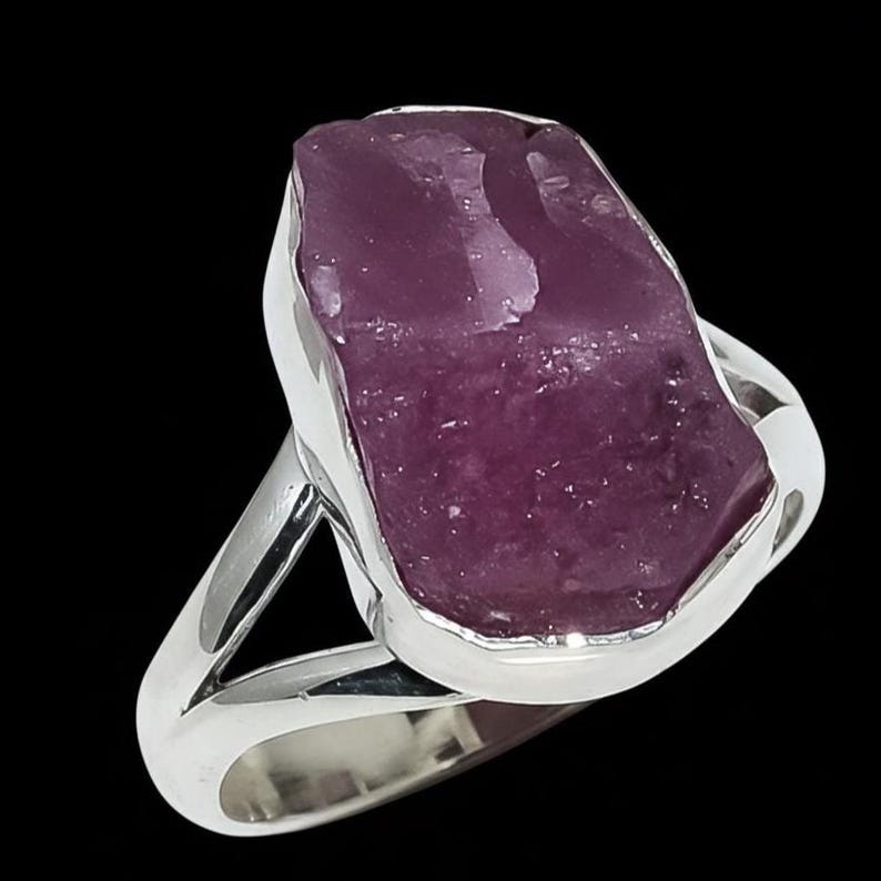 K&ouml;nnte beinhalten: Ein Silberring mit einem gro&szlig;en, roh geschnittenen, purpurrosa Edelstein. Der Stein ist unregelm&auml;&szlig;ig geformt und in einem polierten Silberband gefasst. Der Ring wird vor einem schwarzen Hintergrund gezeigt, um die Farbe und Textur des Edelsteins hervorzuheben.