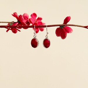 Op de afbeelding: Een paar zilveren oorbellen met rode ovale edelstenen. De oorbellen hangen aan een tak met rode bloemen.
