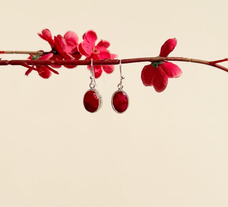 Op de afbeelding: Een paar zilveren oorbellen met rode ovale edelstenen. De oorbellen hangen aan een tak met rode bloemen.