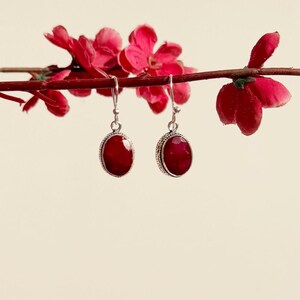 Op de afbeelding: Een paar zilveren oorhangers met ovale rode edelstenen. De oorbellen hangen aan een tak met rode bloesems.