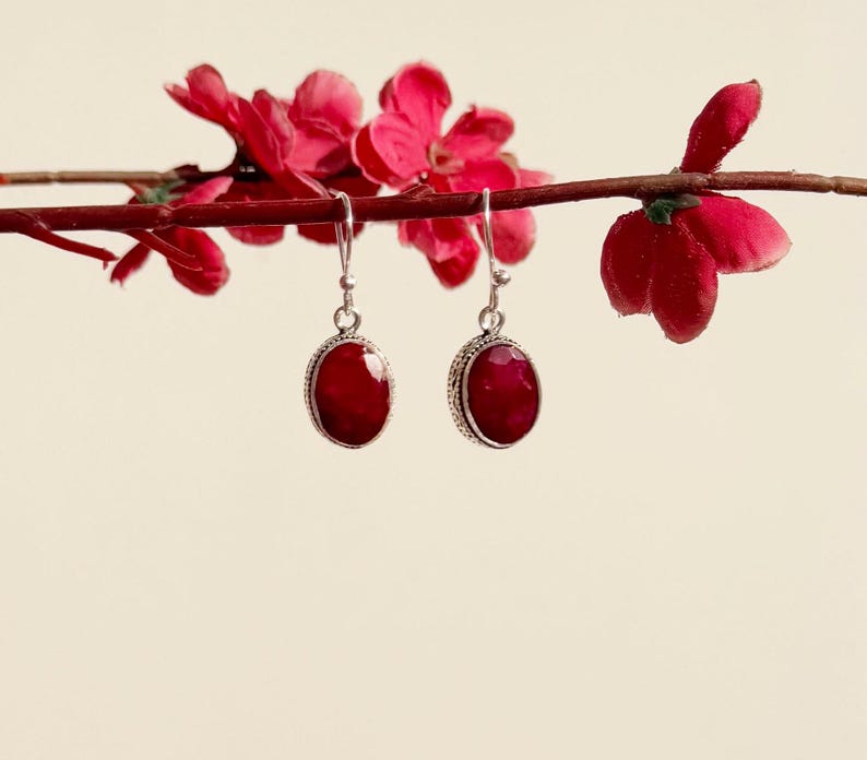 Op de afbeelding: Een paar zilveren oorhangers met ovale rode edelstenen. De oorbellen hangen aan een tak met rode bloesems.