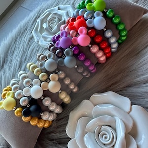 Könnte beinhalten: Ein Set aus bunten Perlenarmbändern mit Mickey-Maus-Anhängern. Die Armbänder sind aus verschiedenen Perlenfarben gefertigt, darunter weiß, schwarz, gelb, gold, braun, grau, lila, rot, rosa, blau und grün.