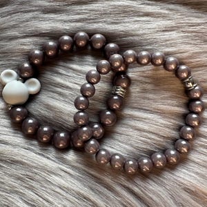 Könnte beinhalten: Zwei graue Perlenarmbänder mit einem weißen Mickey Mouse-Anhänger an einem Armband.