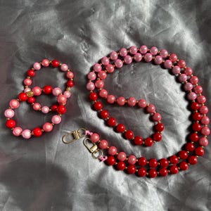 Könnte beinhalten: Ein Set aus Perlen-Schmuck in Rot- und Rosatönen. Das Set umfasst ein Armband, eine Halskette und ein Schlüsselband mit goldfarbenen Verschlüssen. Die Perlen sind rund und scheinen aus poliertem Material zu sein.