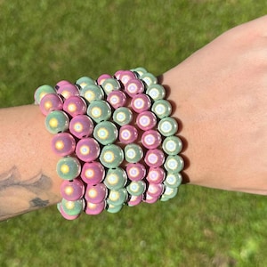 Könnte beinhalten: Eine Sammlung von Armbändern mit Perlen in Rosa- und Grüntönen. Die Armbänder bestehen aus runden Perlen mit schimmerndem Finish. Die Armbänder sind auf einem Handgelenk gestapelt und zeigen die Farbvariationen und Texturen.