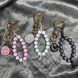 Peut inclure: Trois porte-clés avec des accessoires dorés. Chaque porte-clés a un charme différent : un visage souriant rose, un visage souriant blanc et un visage souriant noir. Chaque porte-clés a également un bracelet de perles rose, blanc et violet.