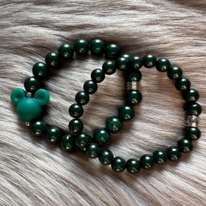 Könnte beinhalten: Zwei dunkelgrüne Perlenarmbänder mit silbernen Akzenten. Ein Armband hat einen grünen Mickey Mouse-Anhänger.