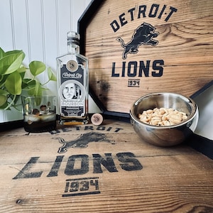 May include: A wooden box with a Detroit Lions logo and the text "Detroit Lions 1934". A bottle of Detroit City Bourbon Whiskey is on the box, along with a glass of whiskey and a bowl of peanuts.