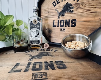 May include: A wooden box with a Detroit Lions logo and the text "Detroit Lions 1934". A bottle of Detroit City Bourbon Whiskey is on the box, along with a glass of whiskey and a bowl of peanuts.