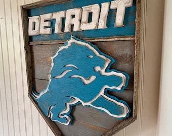 May include: A wooden wall sign featuring the word DETROIT in white block letters above a blue lion silhouette. The sign is constructed from horizontal planks with a weathered finish, framed in rough-hewn wood, and shaped like a home plate.