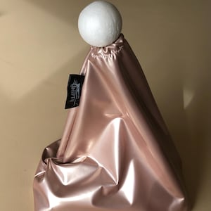 Könnte beinhalten: Eine rosa glänzende Tasche mit einem weißen Ball oben. Die Tasche hat einen Kordelzugverschluss und ein kleines schwarzes Etikett mit weißem Text "SHIFT". Die Tasche besteht aus einem reflektierenden Material.