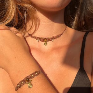 May include: A brown macrame choker necklace and matching bracelet set. The jewelry features gold-colored beads and a central pendant with a spiral design and a round, amber-colored stone. The jewelry is worn on a person.