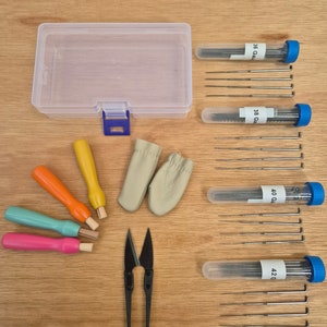 Op de afbeelding: Een set breinaalden in verschillende maten, variërend van 36 gauge tot 42 gauge, in doorzichtige plastic buizen met blauwe dopjes. De set bevat ook een doorzichtige plastic opbergdoos, vier kleurrijke houten breinaalden, twee beige vingerbeschermers en een paar zwarte borduurscharen.